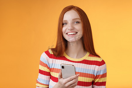 友好热情的年轻红发女孩蓝眼睛，使用智能手机转动摄像头回答，微笑着告诉谁发了消息，站着高兴的橙色背景消息，给男朋友发短信