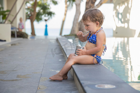 儿童泳摄影照片_坐在室外泳池边用勺子吃冰淇淋酸奶的小女孩