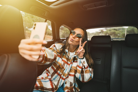 时尚的年轻女子坐在车后座上，用智能手机自拍。