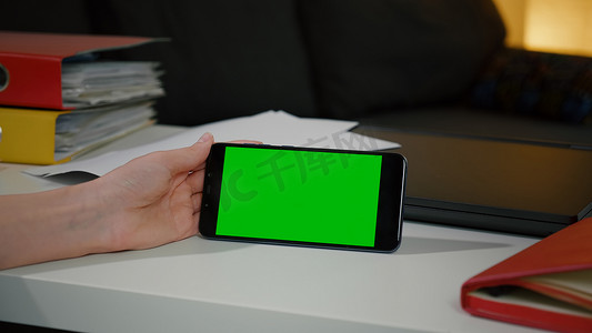 女人坐在办公桌前看着带绿色屏幕色度键显示屏的智能手机。
