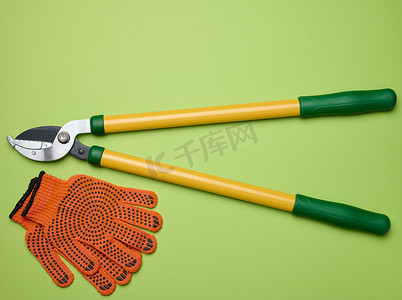 用于修剪树枝的大型花园修剪器和绿纸背景上的橙色纺织手套，顶视图