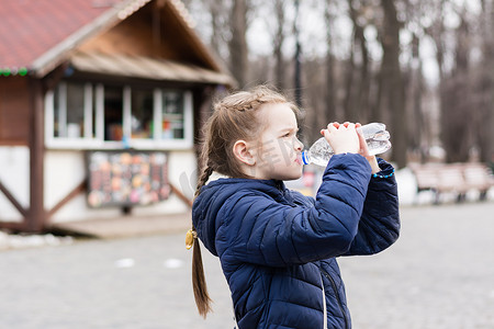 可爱的小女孩在城市公园的餐车背景下喝瓶子里的水。