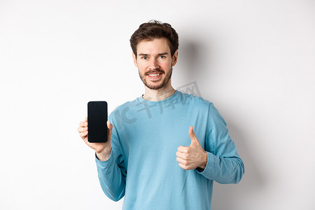 微笑的年轻人展示空的智能手机屏幕和竖起大拇指，称赞好的应用程序，推荐移动应用程序，站在白色背景上