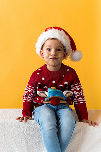 快乐微笑积极快乐积极学龄前小男孩的画像，戴着红色温暖的圣诞老人帽子，在橙色、黄色背景上竖起大拇指。