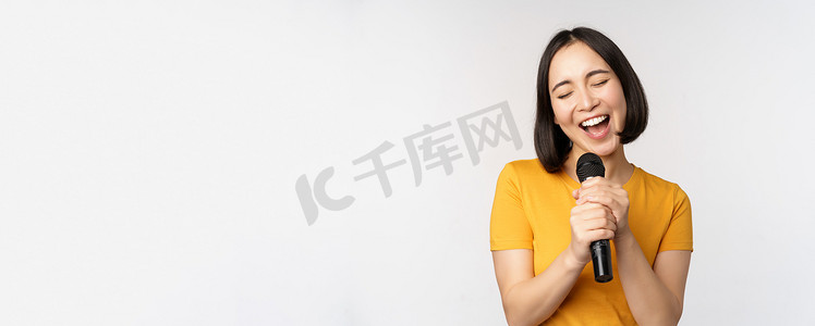 快乐的亚洲女孩唱歌和玩乐，在卡拉 OK 拿着麦克风，站在白色背景下的黄色 T 恤