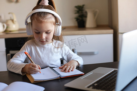 戴耳机的小女孩坐在办公桌前在笔记本上写字在线学习在家做运动，小孩子手写准备隔离作业，上网络课或在室内上课。