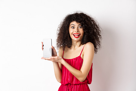 穿着红色连衣裙和化妆的时尚女士，展示手机屏幕并微笑，介绍智能手机应用程序，站在白色背景上