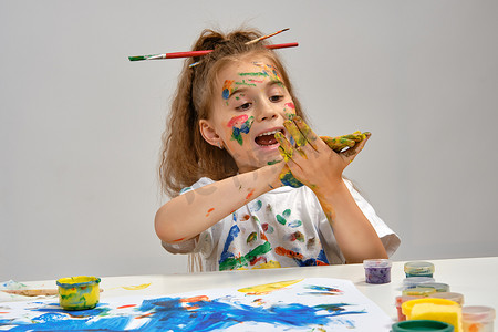 穿着白色 T 恤、头发上梳着刷子的小女孩坐在桌边，手里拿着什么东西和油漆，在上面画画。