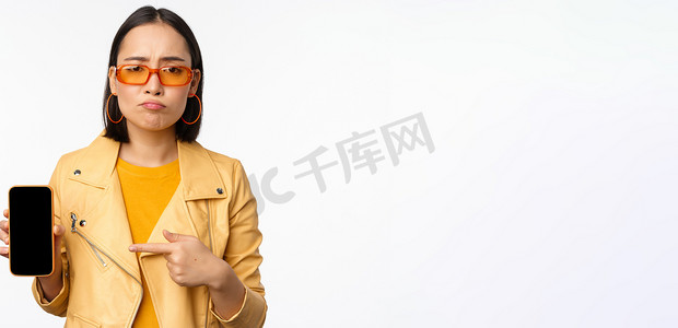 戴着太阳镜的悲伤亚洲女性的肖像，手指指向手机应用程序界面，显示智能手机应用程序，站在白色背景上