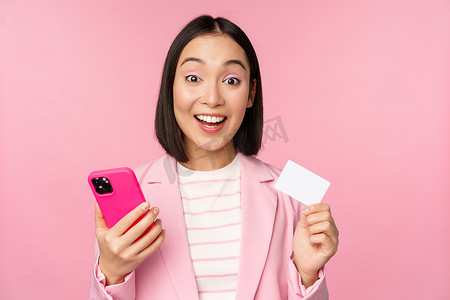 在线支付图片摄影照片_微笑快乐的亚洲女商人展示信用卡、在智能手机应用程序上在线支付、用手机订购、站在粉红色背景下的图片