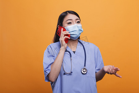 年轻的亚洲护士在用智能手机与病人交谈时戴着面罩和听诊器。