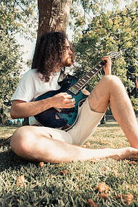 年轻的长发嬉皮士靠着一棵树在公园里微笑着弹吉他。