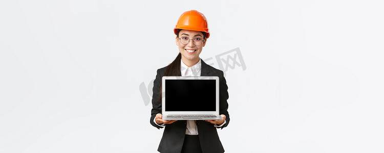 带安全头盔摄影照片_微笑的亚洲专业女工程师向投资者或客户介绍施工计划，站在安全头盔和西装上，带着愉快的微笑、白色背景展示笔记本电脑屏幕