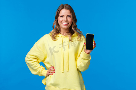 穿黄色连帽衫的腰上肖像漂亮金发女孩推广智能手机应用程序，展示手机显示屏和微笑相机，推荐订阅或下载应用程序