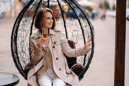 一个穿着灰色外套的快乐时尚女孩坐在外面的扶手椅上喝着饮料