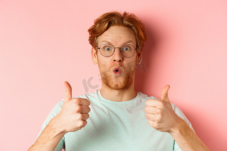 戴着眼镜和 T 恤的快乐红发男人的脸，竖起大拇指，看起来很兴奋，赞同和赞美酷炫的促销活动，站在粉红色背景上