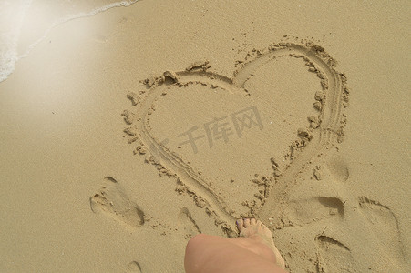 沙画女人摄影照片_你好夏天，站在沙滩上，女人的脚在湿沙上画出一个心形标志。