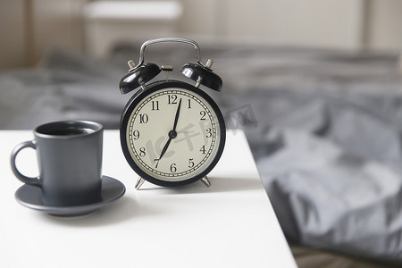 图片闹钟，床前的白色床头柜上放着一杯黑咖啡，床前配有灰色床单。