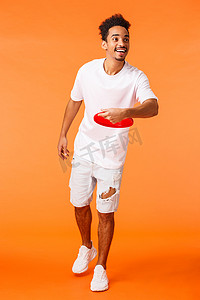 全长垂直拍摄欢快的非洲裔美国可爱男人扔飞盘，开心地笑着，在户外和朋友玩游戏，享受假期，假期休闲，橙色背景