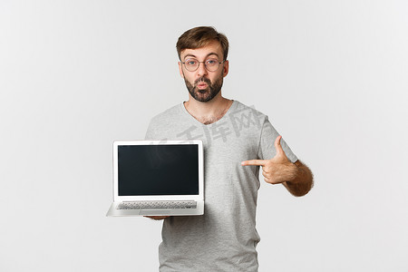 留着胡子、穿着灰色 T 恤和眼镜的帅气白种人指着笔记本电脑屏幕，在网上展示一些东西，站在白色背景上