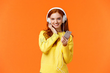 冬天的音乐背景摄影照片_穿着黄色毛衣的时髦漂亮红发女性，听音乐白色耳机，触摸耳机和手持智能手机，听音乐，享受良好的耳塞质量，微笑，橙色背景