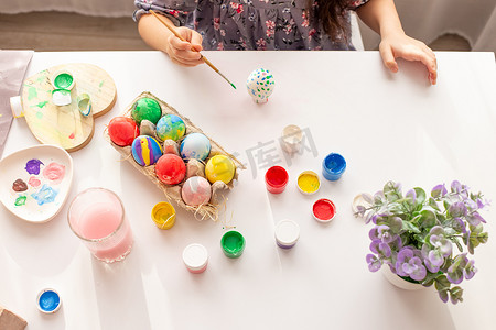 一个小女孩的手上拿着一个彩绘的鸡蛋，放在一张白色的桌子上，托盘里放着彩色的鸡蛋、刷子和油漆。