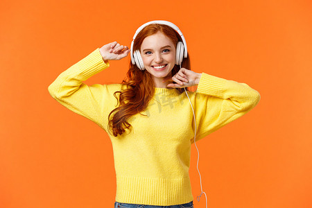 快乐而无忧无虑兴奋的红发时髦女孩喜欢她的新耳机，举起双手跳舞，微笑着享受橙色背景下的音乐，用最喜欢的歌曲提振情绪