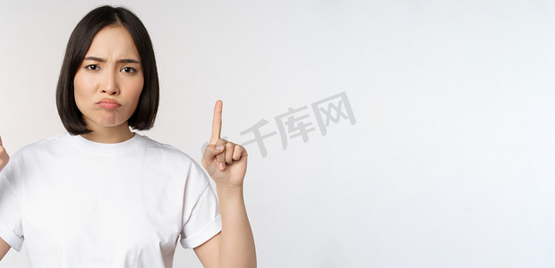 悲伤而失望的亚洲年轻女性竖起手指，展示带有不安表情、白色工作室背景的广告