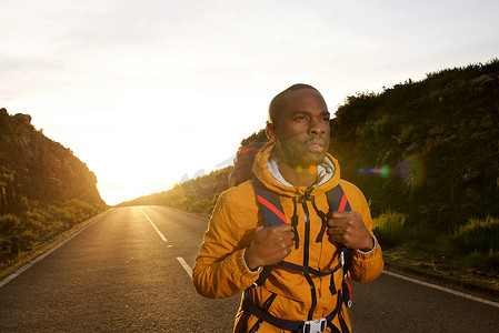 背包的人走在路上摄影照片_日落时背着背包走在路上的年轻非洲裔美国徒步旅行者