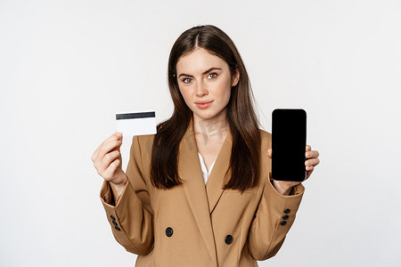 女商人展示手机屏幕和信用卡，展示在智能手机上的应用，穿着西装站在白色背景中