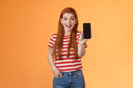 app展示摄影照片_兴奋可爱的外向红发女性印象深刻的展示应用程序，持有智能手机介绍小工具功能，微笑惊讶，看起来热情像应用程序，吹牛游戏分数，橙色背景