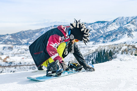 戴着范妮帽的年轻男子单板滑雪者坐在雪地里，在下山前把扣子固定在滑雪板上