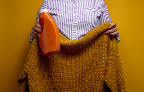 空白的女士衬衫摄影照片_一位身穿白色条纹衬衫的女士拿着一个装有液体洗涤凝胶的橙色塑料瓶和一件针织毛衣。