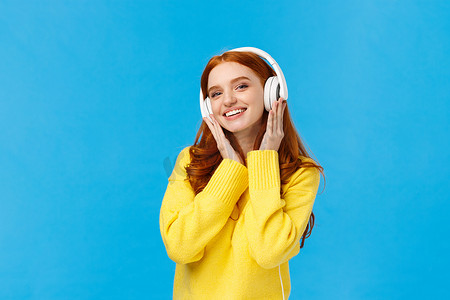 穿着黄色毛衣的时髦迷人、无忧无虑的红头发女人戴着耳机听音乐，快乐地微笑着倾斜头，收到新耳机作为圣诞礼物，站在蓝色背景