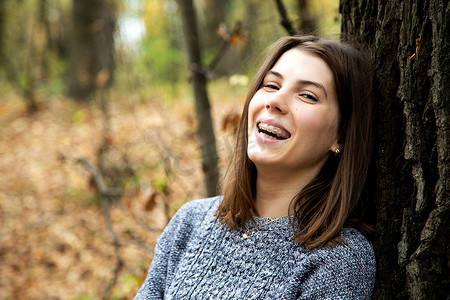 身穿灰色毛衣、牙齿上戴着牙套的年轻美女坐在秋天的森林里，靠近一棵大树，甜美地微笑