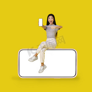 亚洲年轻女孩拿着智能手机坐在巨大的智能手机上，黑色屏幕隔离在黄色背景上。