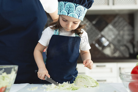有爱心的母亲教小女儿在厨房做沙拉，年轻的妈妈和可爱的女孩穿着围裙，在台面上用刀切菜，站在家里的厨房里。