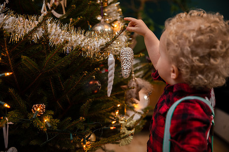 一个两岁的金色卷发男孩在圣诞树上挂着一个小玩意儿。