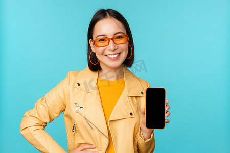 戴着时尚太阳镜的年轻时尚亚洲女模特的形象，显示手机屏幕，智能手机界面上的应用程序，站在蓝色背景上