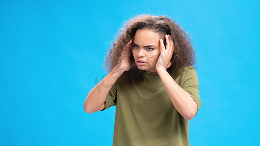 压力大或不开心的非洲裔美国女孩穿着蓝色背景中突显的橄榄色 T 恤，摸着头疼。