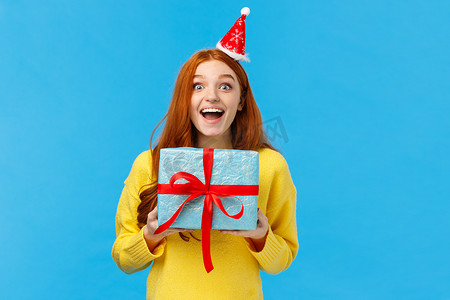 新年蓝色红色摄影照片_兴奋的女孩喜欢圣诞假期和收到礼物，拿着可爱的新年礼物，开心地笑着，开心地笑着，戴着圣诞帽参加派对，站在蓝色背景上乐观