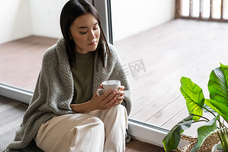 年轻悲伤的亚洲女人坐在阳台附近的地板上，看着茶杯若有所思