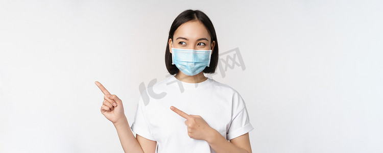 戴着医用面罩的韩国年轻女性手指向左，看着标志，展示广告或横幅，站在白色背景上
