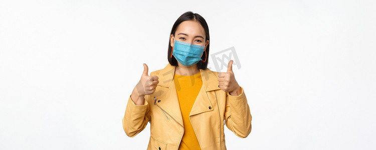 戴着医用口罩的亚洲女性微笑着竖起大拇指，支持佩戴来自covid-19、冠状病毒大流行、白色背景的防护装备