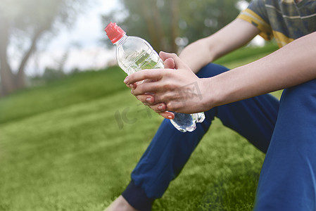 清晨跑步摄影照片_年轻的白人男子跑步者在清晨运动后放松地拿着饮用水瓶，坐在公园户外的草地上，锻炼身体和健康理念。