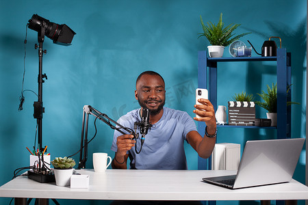 音频博客摄影照片_坐在视频博客工作室里的视频博主拿着麦克风用智能手机自拍