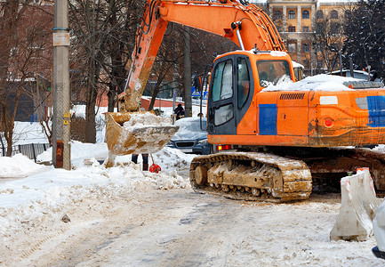 雪街道摄影照片_带有金属铲斗的大型履带式挖掘机正在清理城市街道上堆积如山的积雪。