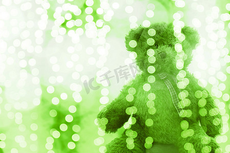焦点效应摄影照片_泰迪熊娃娃在照明线散景绿色白色明亮圣诞节或新年快乐背景，熊在闪光绿色背景模糊散景明亮（选择性焦点）