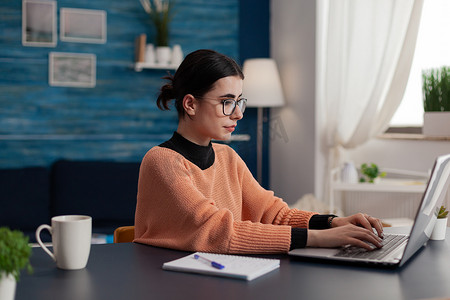 戴眼镜的学生坐在家里的办公桌前在笔记本电脑上打字。
