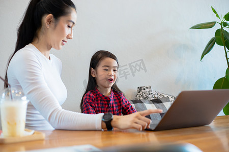 学校放假摄影照片_亚洲母亲在学校放假期间在家教女儿读书并使用笔记本和技术进行在线学习。